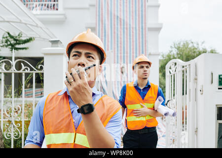 Serious frowning Vietnamese construction engineer talking on walkie-talkie when he and his coworker leaving building they were working in Stock Photo