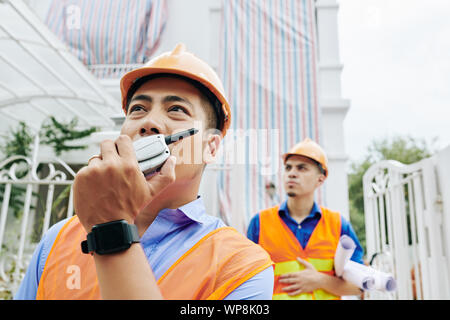 Serious Asian contractor controlling work at construction site by talking on walkie-talkie with colleagues Stock Photo