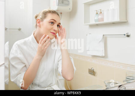 Pretty young woman standing in front of mirror and doing sculpting face massage after shower Stock Photo