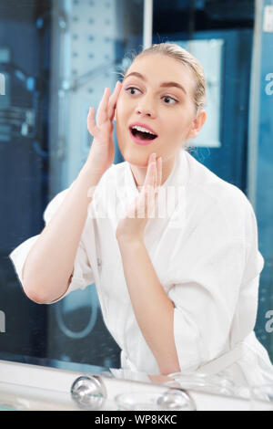 Portrait of excited young woman in bath robe applying miraculous moisturizing and firming lotion on her face Stock Photo