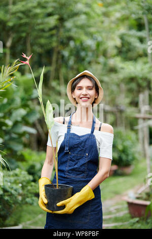 Smiling female garden worker in denim uniform carrying pot with blooming flower Stock Photo