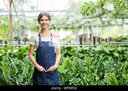 Positive pretty young woman standing in front of greenhouse full of various plants and flowers and smiling at camera Stock Photo