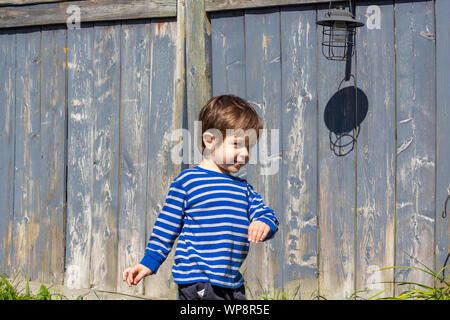 An adorable little boy, about two years old, moves his arms as he walks alongside a fence. Stock Photo
