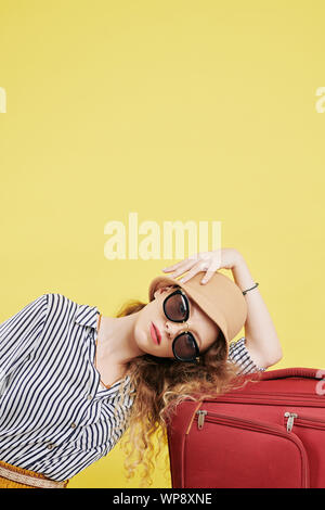 Beautiful young woman in big sunglasses resting her head on big packed suitcase and looking at camera, isolated on yellow background Stock Photo