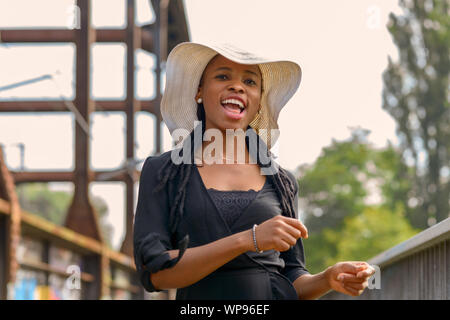 Laughing vivacious young African woman wearing braided hair extensions and a stylish wide brimmed straw hat in a close up outdoor portrait Stock Photo