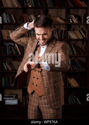 a young man with a beard in a brown suit with a smile sits on his watch and holds his head while standing in the library. A businessman smiles and keeps track of time Stock Photo