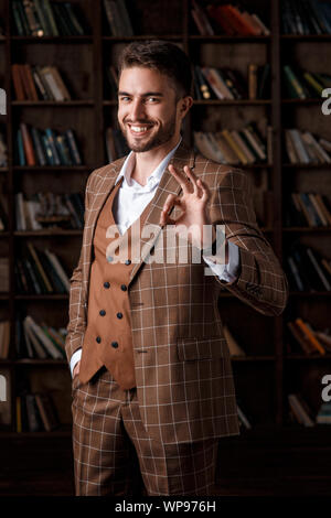 young man with a beard in a brown suit shows ok gesture standing in the library. The businessman is smiling and pleased with the result Stock Photo