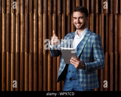 a man in a suit with a tablet in his hands shows a class gesture. businessman in a blue checked suit in a loft studio. Stock Photo