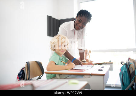 Pleasant dark-skinned teacher standing near pupil Stock Photo