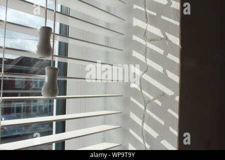 Light coming through white venetian blinds with the strings, and plain fairy lights Stock Photo