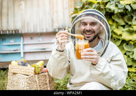 Portrait of a cheerful beekeeper in protective uniform with honey products and sweet fruits on the apiary with beehives on the background Stock Photo