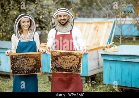 Portrait of a male and female beekeepers in protective hats and aprons holding honeycombs on the apiary with wooden beehives on the background Stock Photo