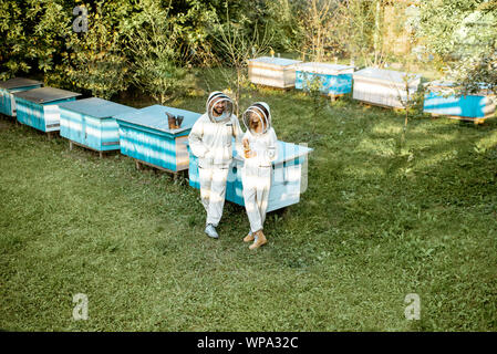 Two beekeepers in protective uniform standing together near the wooden beehives on a small traditional apiary. Wide view from above Stock Photo