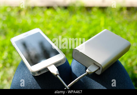 White smartphone charged by power bank on the knees on the background of green grass. Stay connected while traveling or camping Stock Photo