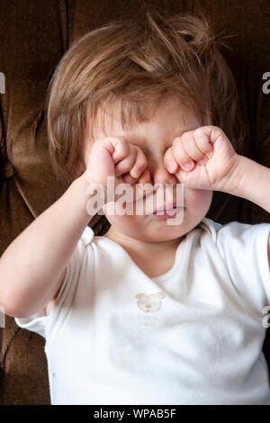 Caucasian child, boy, 2-3 year old, sitting in white romper suit on brown settee, rubbing both eyes with fists after waking up. Close up. Stock Photo