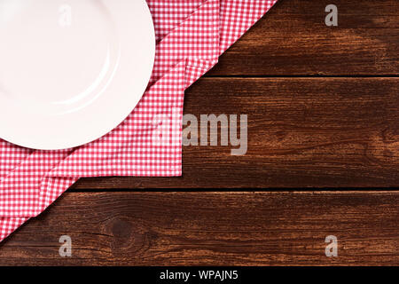 Flat lay of old wooden background with red checkered dishcloth, round white plate and space for text. Stock Photo