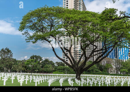 Manila, Philippines - March 5, 2019: American Cemetery and Memorial park. Green field filled with white crosses. Trees and bushes in front. High rise Stock Photo