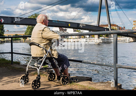 Old Man sitting on his wheeled walker, Old Man sitting on his Rollator, Old Man sitting on Riverside, Bridge and Clouds in Background Stock Photo