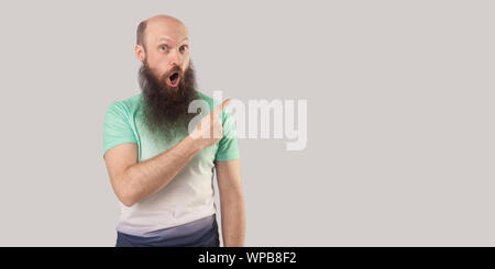 Portrait of surprised middle aged bald man with long beard in light t-shirt standing, looking with shocked face and pointing at wall empty copyspace. Stock Photo