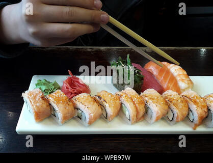 Set with philadelphia rolls, sushi, ginger and wasabi on square white plate and on wooden table surface. Boy's hand is ready to take a roll with chops Stock Photo