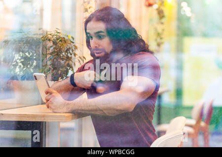 Portrait through the window of unbelievable handsome young adult muscular man with curly long hair sitting on cafe, holding phone with shocked face po Stock Photo