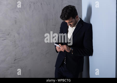 A serious handsome young businessman in a black suit using a smartphone in front of a grey wall in a studio. Stock Photo