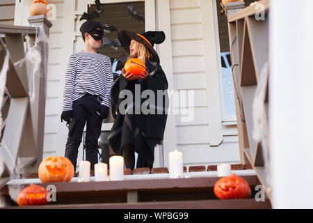 Low angle portrait of two children, boy and girl, standing on porch of decorated house while trick or treating on Halloween, copy space Stock Photo