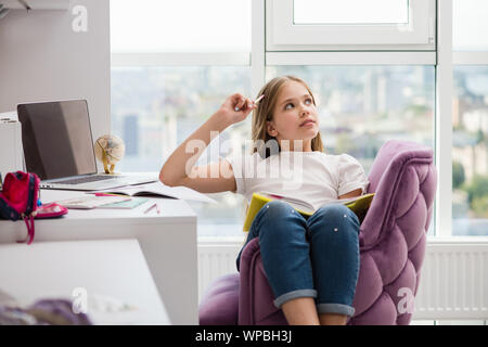 Joyful girl holding pencil near head looking up thinking dreaming. School supplies and laptop in background. Stock Photo