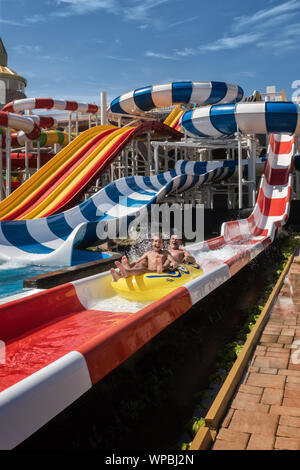 Family in water splash after riding down the water park steep structure, sitting together on yellow inflatable ring.  Father and son are enjoying aqua Stock Photo