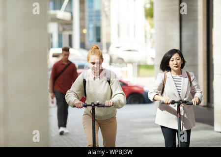 Portrait of two women riding electric scooters while commuting in urban city, copy space Stock Photo