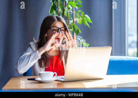 Portrait of angry brunette young woman in glasses sitting, looking at her laptop screen on video call and screaming to share something important. indo Stock Photo