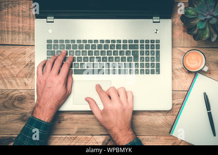 Office work top view with hands typing on a laptop. Rustic wooden desk with computer, plant, coffee and notebook. Modern workspace concepts. Stock Photo