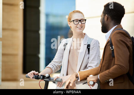 Waist up portrait of modern young couple riding electric scooters in city street, focus on red haired woman smiling cheerfully, copy space Stock Photo