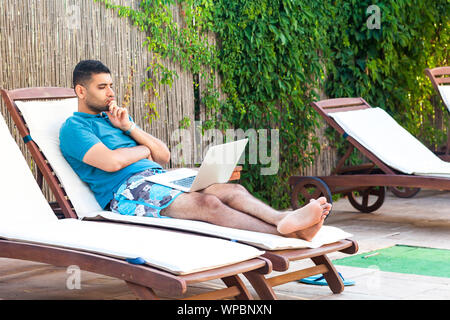 Portrait of serious handsome bearded young adult freelancer man in blue t-shirt and shorts lying on cozy deck chair with laptop on poolside, working o Stock Photo