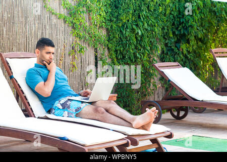Portrait of clever handsome bearded young adult freelancer man in blue t-shirt and shorts lying on cozy deck chair with laptop on poolside, working on Stock Photo