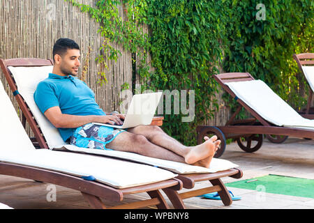 Portrait of pleasure handsome bearded young adult freelancer man in blue t-shirt and shorts lying on cozy daybed with laptop on poolside and working o Stock Photo