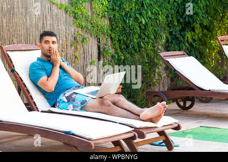 Portrait of suprised bearded young adult freelancer man in blue t-shirt and shorts lying on deckchair on cozy poolside and looking at camera with big Stock Photo