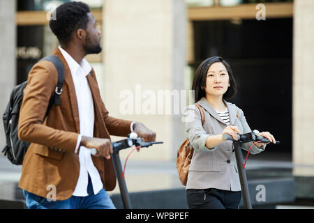 Waist up portrait of Asian woman riding electric scooter in street together with African man, copy space Stock Photo