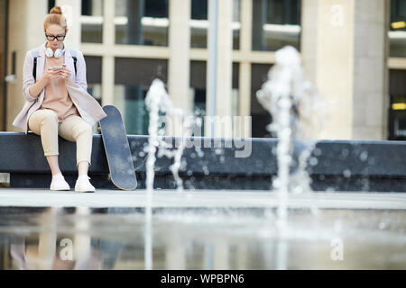 Full length portrait of contemporary young woman using smartphone sitting on bench in urban setting, copy space