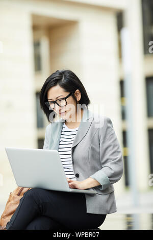 Portrait of modern Asian woman using laptop outdoors sitting on bench in urban setting and looking at screen, copy space Stock Photo
