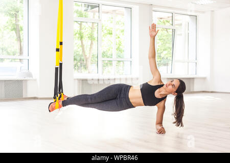 Portrait of young attractive muscular woman in black top and leggings standing on perfect side plank position use fitness straps, doing push ups while Stock Photo