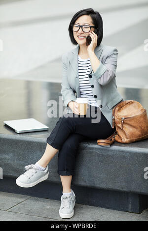 Full length portrait of modern Asian woman speaking by smartphone outdoors and smiling happily, scene in urban setting Stock Photo