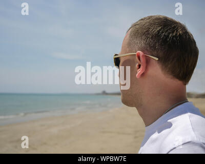 Young attractive man in sunglasses stands alone near the sea and looks at the side of the horizon Stock Photo