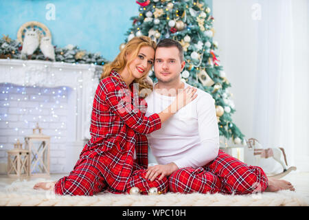 Loving couple husband and wife in plaid pajamas are sitting near the New Year tree by the fireplace and hugging. Love and a happy family Stock Photo