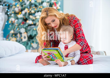 Mom and son in checkered pajamas are sitting on the bed and reading a book of New Year's tales together Stock Photo