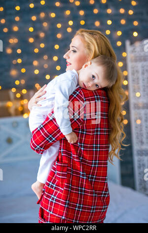 A beautiful young mother in checkered pajamas holds a little son in her arms. Caring and motherly love Stock Photo
