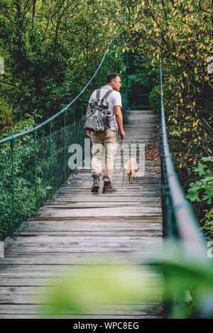 Hiker with small yellow dog walking over wooden suspension bridge in the forest Stock Photo