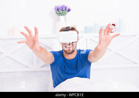 Using computer technology to create a simulated environment. Happy guy wearing VR headset for computer gaming. Handsome man playing virtual computer games in bed. Gaming computer. Stock Photo