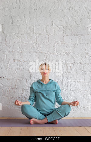 Blond girl in blue sportswear sits on a rug on the floor in a lotus position Stock Photo