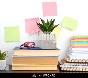stack of spiral notebooks and colored stickers, next to a ceramic pot with a flower on a black table, white wall Stock Photo
